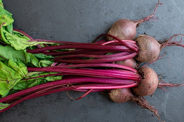 Pile of raw beets with reddish brown roots, bright red stems and green leaves